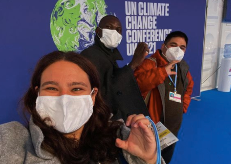 Three people wearing white masks covering their mask make a C shape with their hands. They are standing in front of a sign that reads "UN Climate Change Conference"