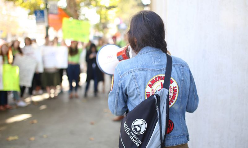 A young leader speaks into a bullhorn with their back turned to the camera. Protestors can be seen in the background.